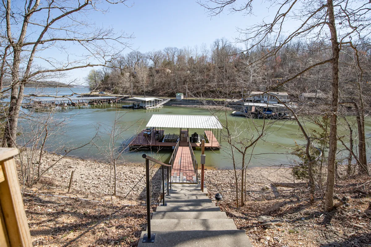 A wide shot of a staircase leading down to the water, with docks extending out onto the lake. Boats can be seen moored at the docks, and the area is surrounded by trees, giving a sense of privacy and access to nature. The water's surface is calm, perfect for swimming or launching a kayak.