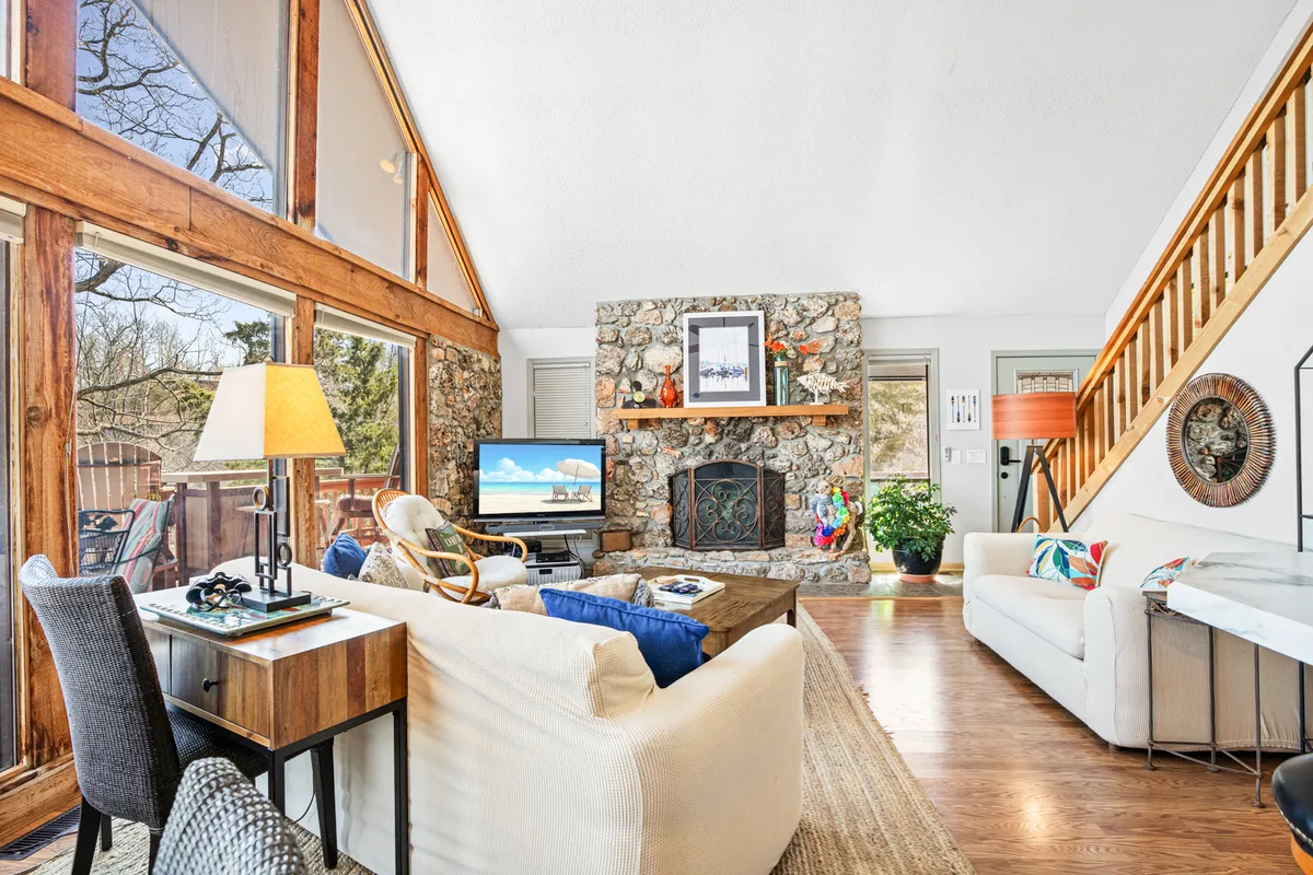 This image features a spacious living room with a large stone fireplace, expansive floor-to-ceiling windows overlooking the lake, a comfortable seating area, and a smart television. The arrangement is perfect for family gatherings, movie nights, and enjoying the lake views from indoors.