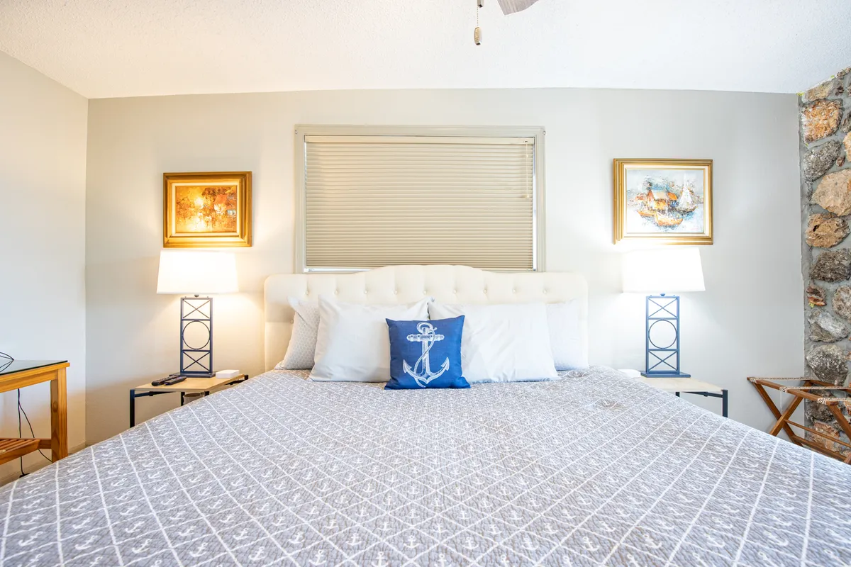 A tranquil bedroom featuring a bed with decorative pillows, framed artwork above the headboard, matching nightstands with lamps, and a textured stone wall adding warmth and character. Soft lighting and subtle decor contribute to a serene and restful atmosphere.