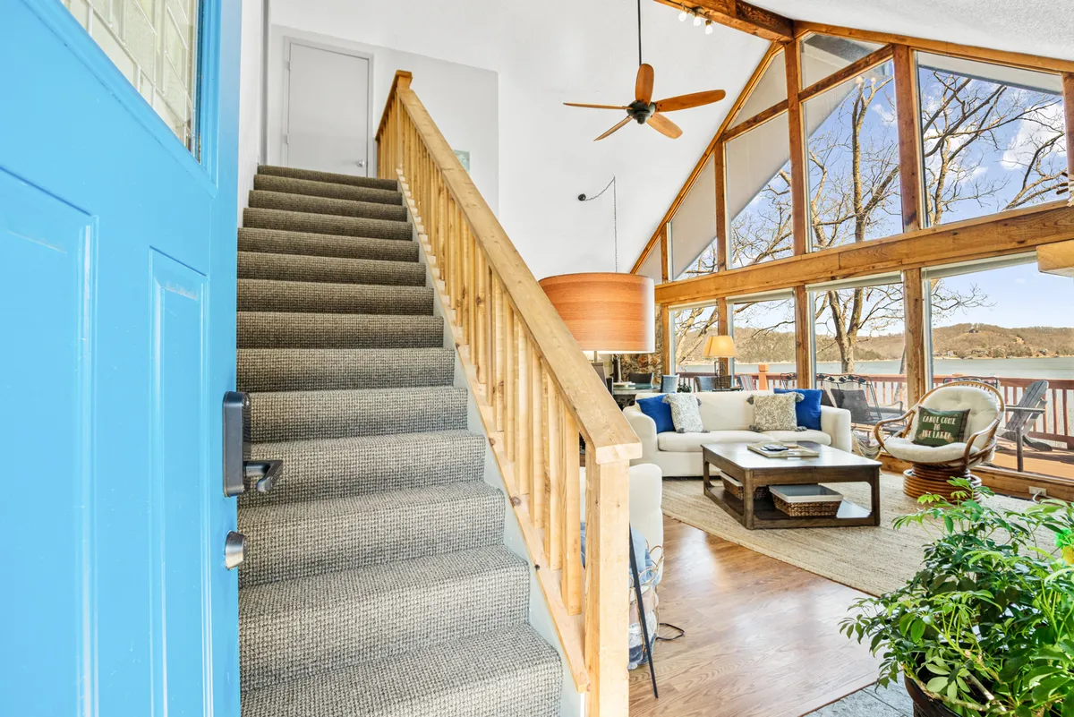 A welcoming living area with a blue front door, comfortable seating, large windows overlooking the water, a decorative lamp, and a staircase. The room is bathed in natural light, providing a cozy yet open atmosphere.