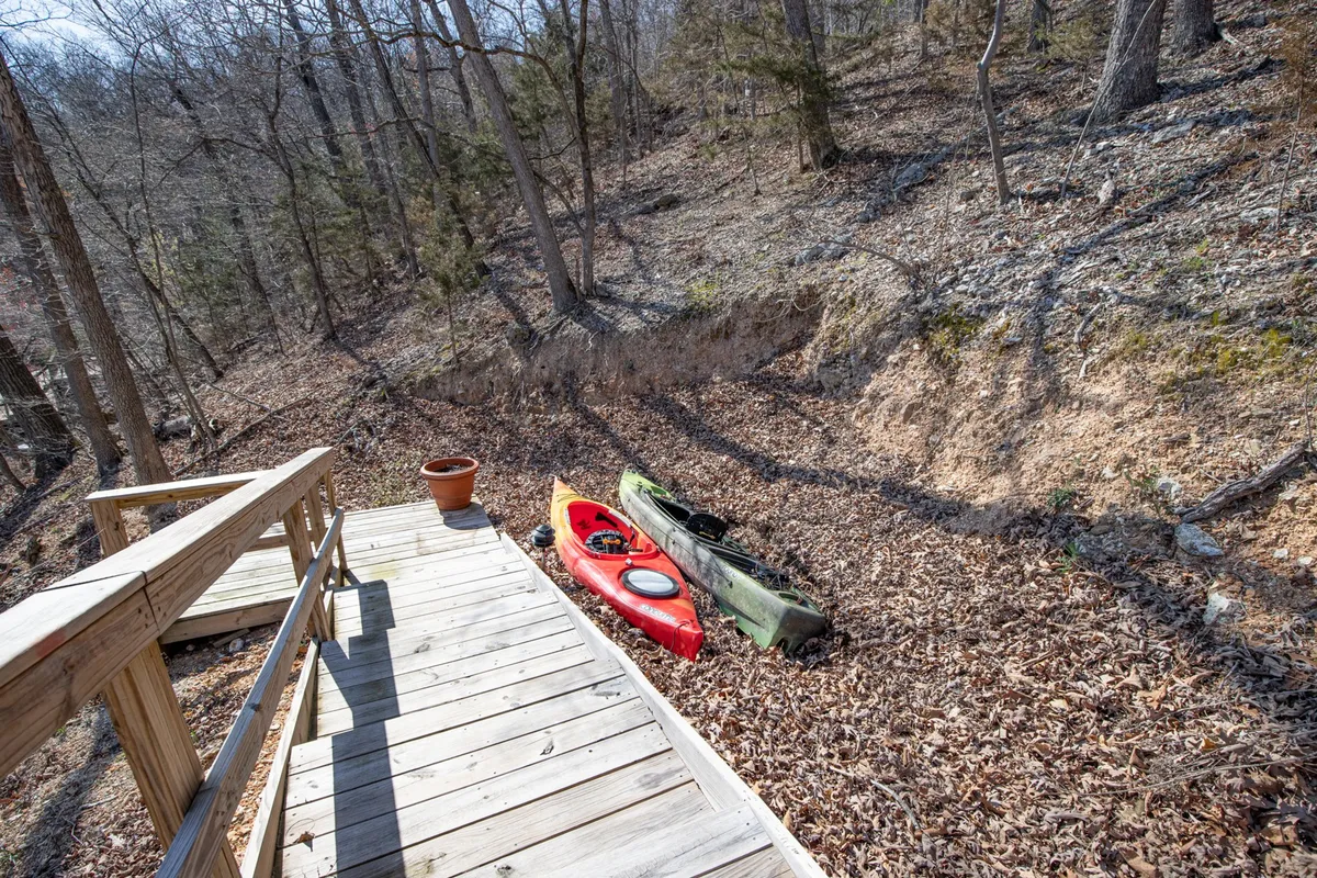 A wooden pathway leads toward the lake, where two kayaks (one red, one green) are ready for adventure. The scene is surrounded by natural greenery, with a flower pot adding a pop of color. The ground is covered with leaves, suggesting a woodland setting ideal for nature walks.
