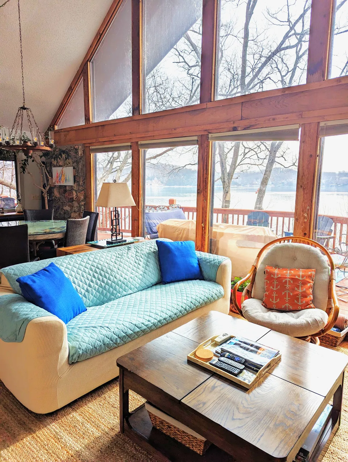 A sunlit living space with large windows overlooking the lake, a comfortable sofa accented with blue cushions, and a wooden coffee table at the center. The area is inviting and ideal for relaxing, reading, or gathering for board games while soaking in the lake view.