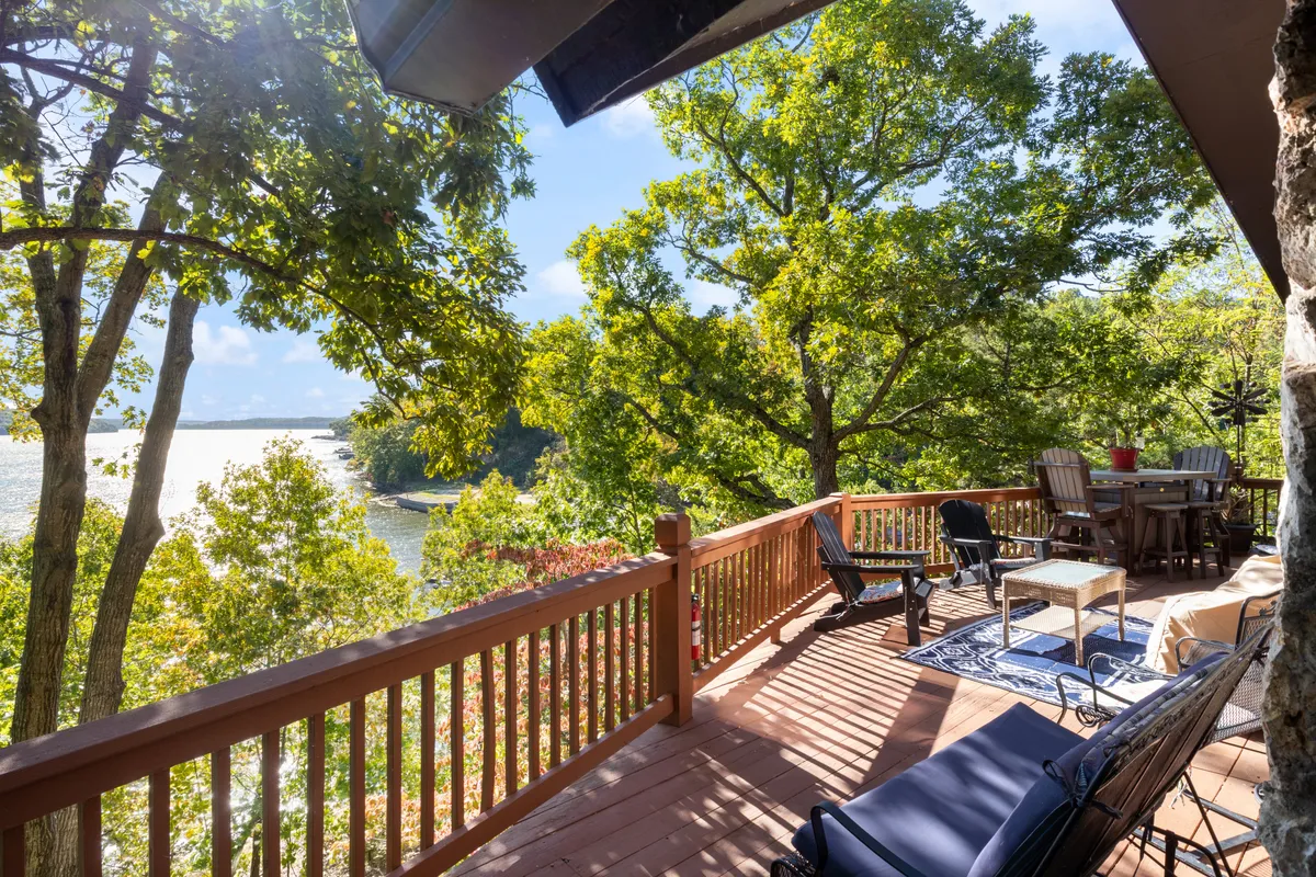 An inviting outdoor deck with a railing that frames a beautiful view of the water. A comfortable seating arrangement and a table with chairs create an ideal space for relaxing, entertaining, or simply soaking up the lakeside atmosphere. Lush greenery surrounds the area, adding to the tranquil ambiance.