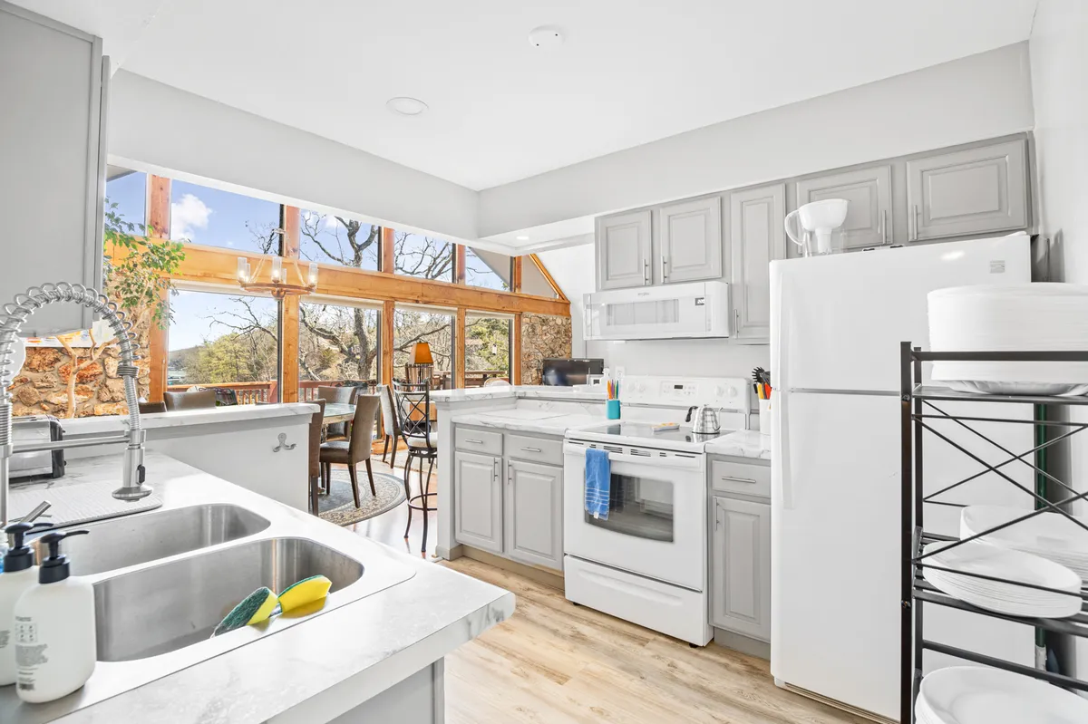 This image features a modern kitchen with large windows flooding the room with natural light, modern appliances, and a welcoming wooden dining set. The kitchen island serves as a focal point for both cooking and gathering. The windows offer partial views toward the outdoors, connecting the kitchen to the scenic lakeside setting.