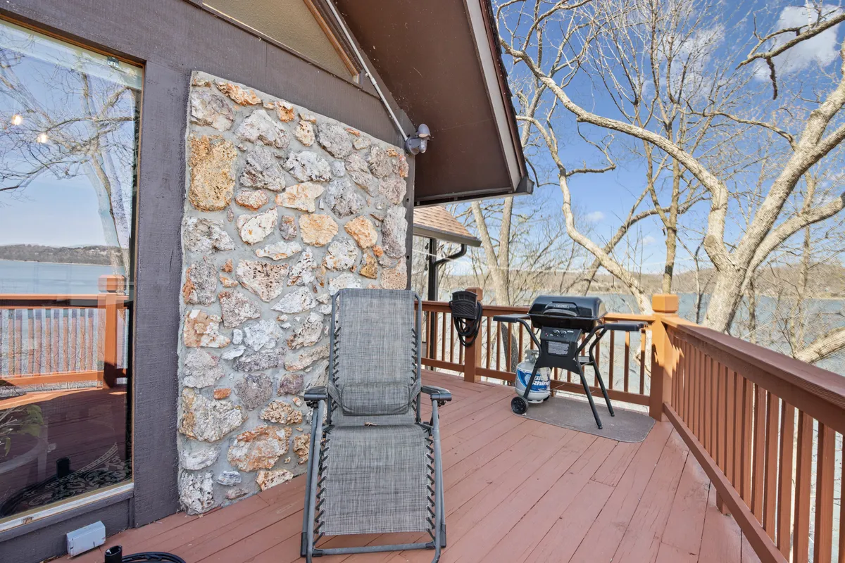 A cozy exterior scene featuring a wooden deck with a grill, stone wall, and an empty chair overlooking a beautiful lake view. The deck is spacious and inviting, ideal for outdoor activities and relaxation.