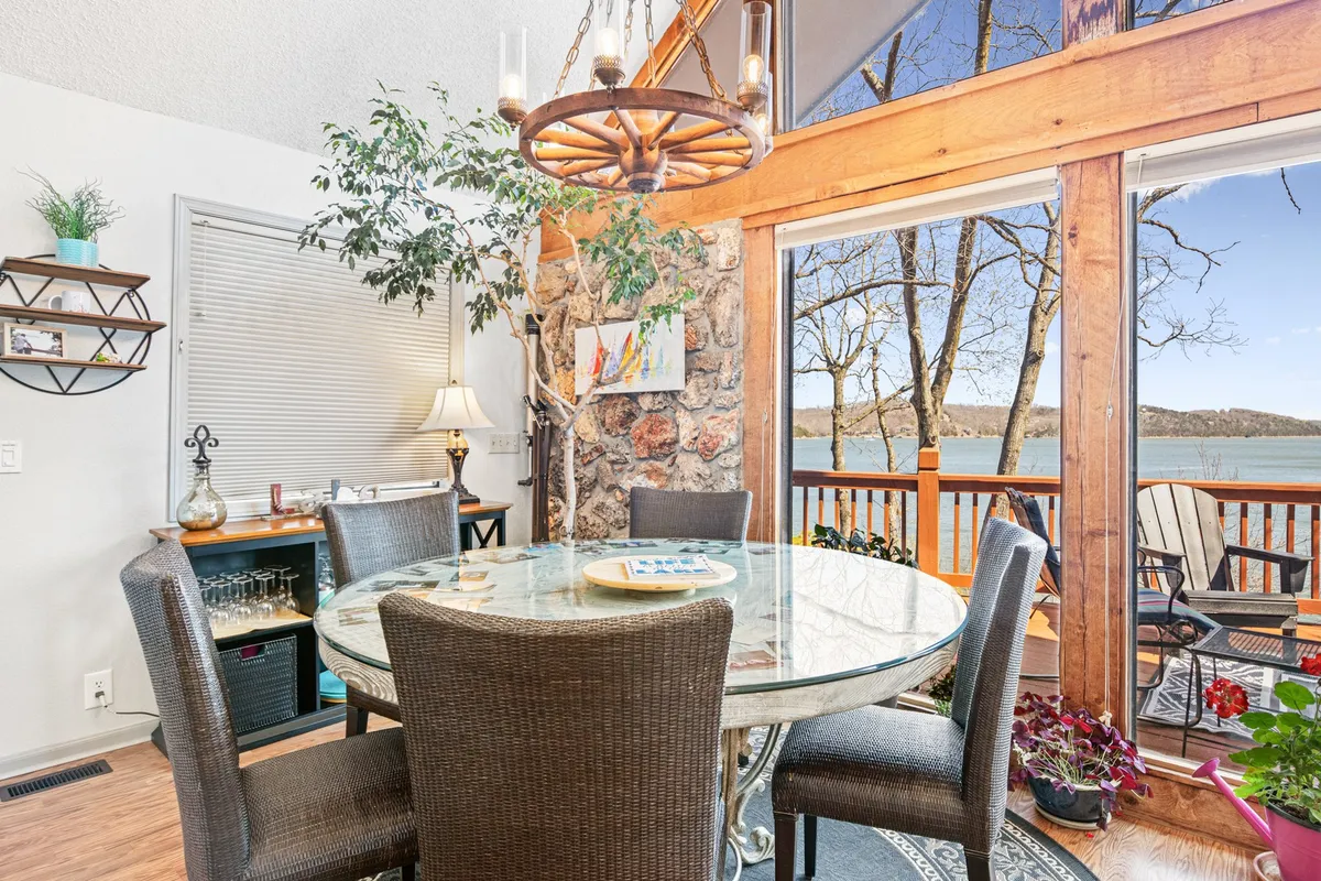 A round dining table set beside large windows overlooking the lake, accented by a rustic stone wall and a central chandelier. The abundance of natural light and open views create a memorable setting for family meals and gatherings.