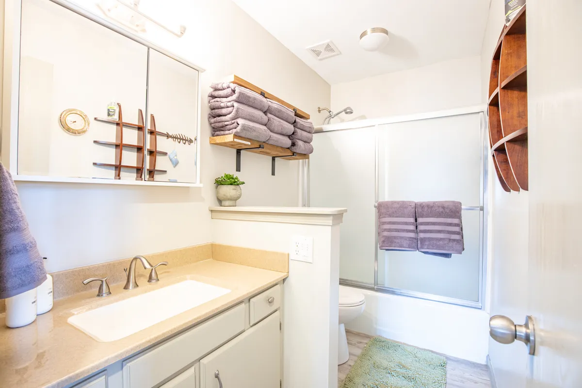 A bright bathroom with a double sink vanity, mirrored cabinet, open shelving with towels, a green rug adding a pop of color, and a glass shower stall. The space feels inviting and thoughtfully designed for relaxation and self-care.