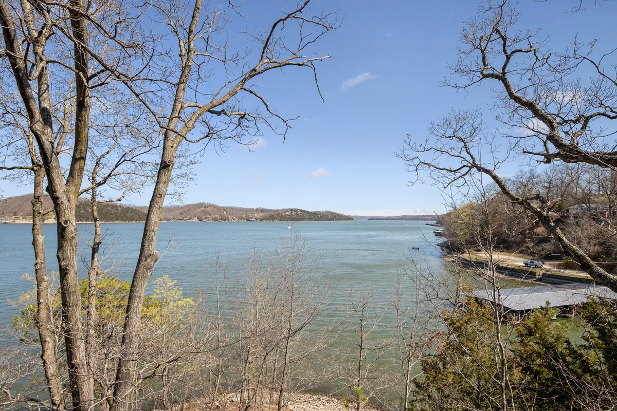 A wide, serene view of the calm lake, distant hills, and trees reflected on the water's surface. The peaceful scene captures the expansive nature of the Lake of the Ozarks and hints at the excitement of watching boat races or fireworks from afar, all while emphasizing the natural beauty and tranquility of the setting.