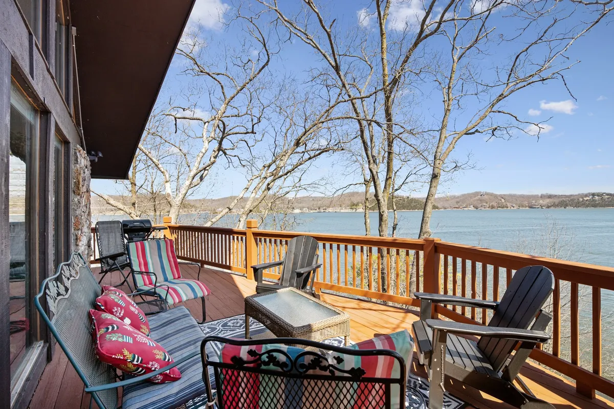 An outdoor wooden deck with comfortable seating and decorative pillows overlooks the tranquil lake. The vantage point showcases the seamless indoor-outdoor flow and provides the perfect spot for watching sunrises or sunsets over the water.