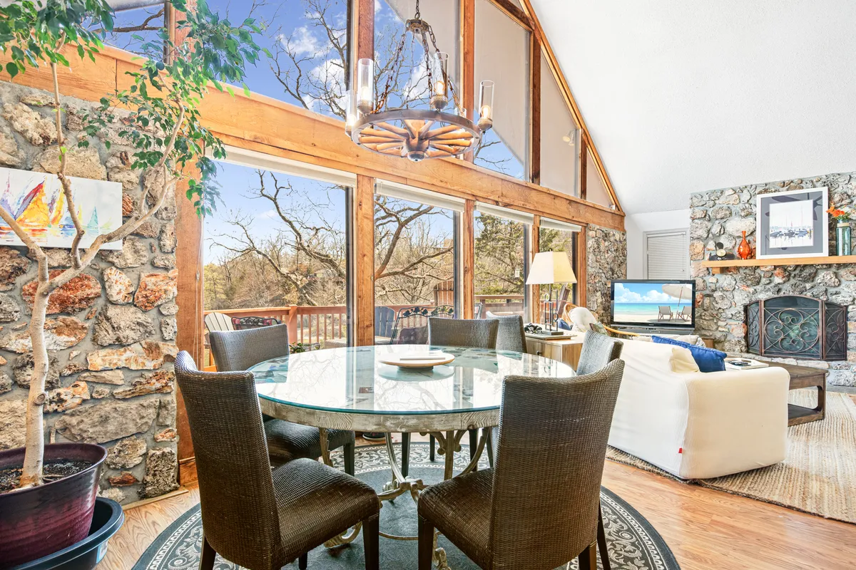 A spacious, sunlit dining area anchors the open-concept living space, framed by expansive windows that reveal tranquil lake views. A grand stone fireplace adds a cozy focal point, while stylish artwork brings personality and warmth. The setting invites guests to gather for meals, games, or fireside chats, all while soaking in the beauty of the outdoors just beyond.