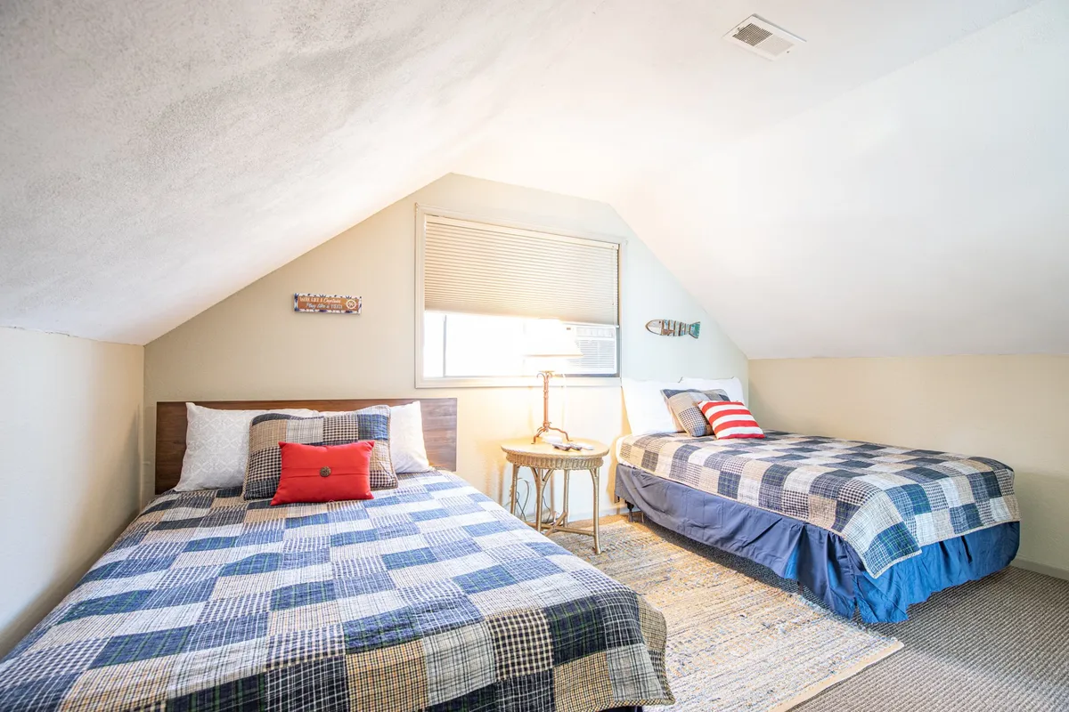 A comfortable, homey bedroom with two beds adorned in patterned quilts, a small side table with a lamp between them, and natural light streaming through a window. The space is functional and flexible, ideal for families or groups needing multiple beds.